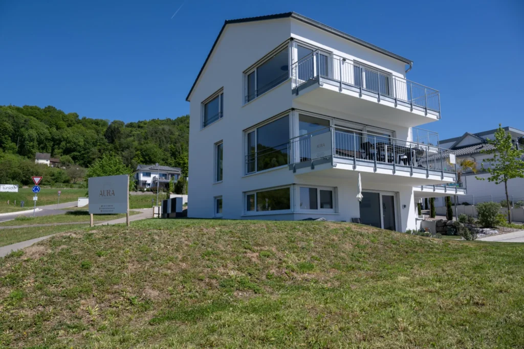 ferienhaus-aura-aussenansicht-frontansicht-schild Frontansicht des Ferienhaus Aura - modernes weisses Ferienhaus mit AURA-Schild, Balkonen und bewaldeten Huegeln