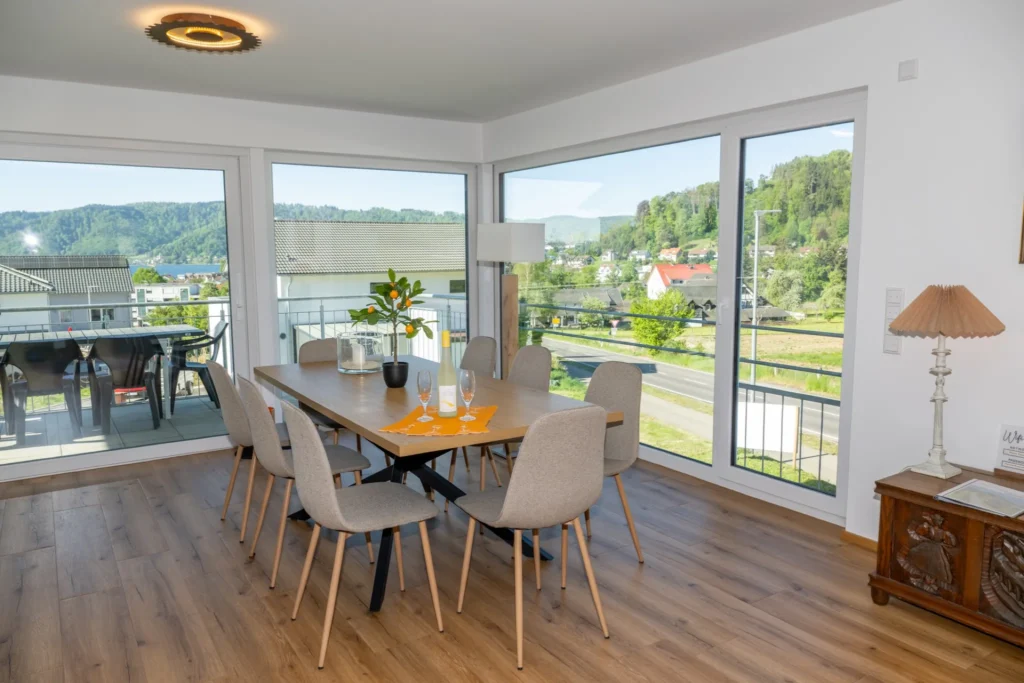 ferienhaus-aura-essbereich-eckfenster-panorama Essbereich im Ferienhaus Aura mit 8-Personen-Esstisch, Eck-Panoramafenstern und Blick auf See und Berge