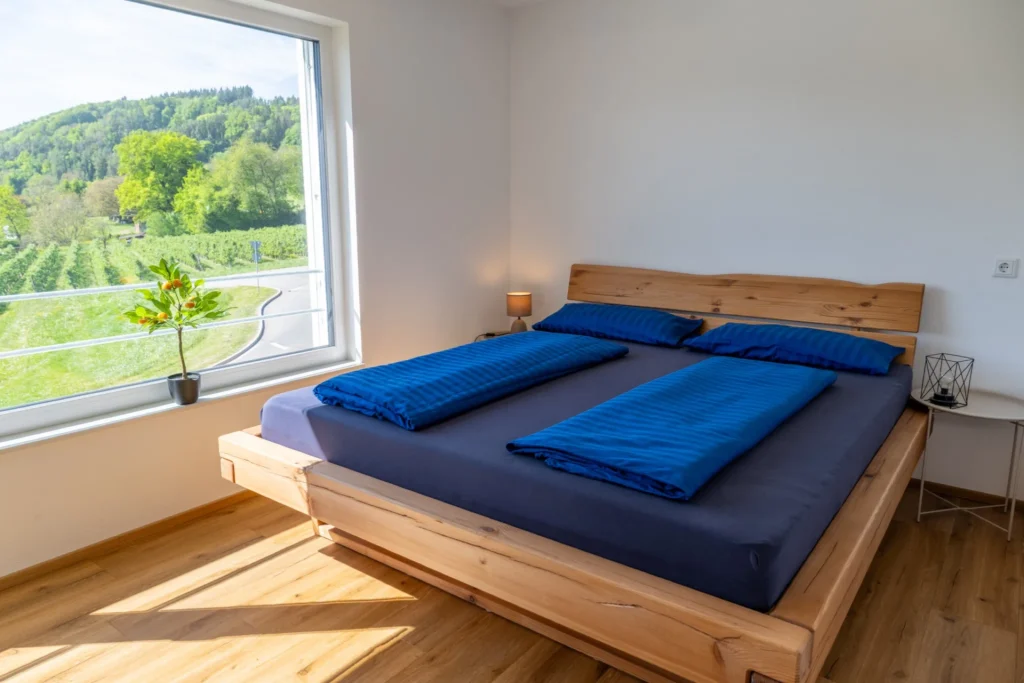 ferienhaus-aura-schlafzimmer-doppelbett-bergblick Schlafzimmer im Ferienhaus Aura mit Eichen-Doppelbett, blauer Bettwaesche und Fenster mit Blick auf Weinberge und Huegel