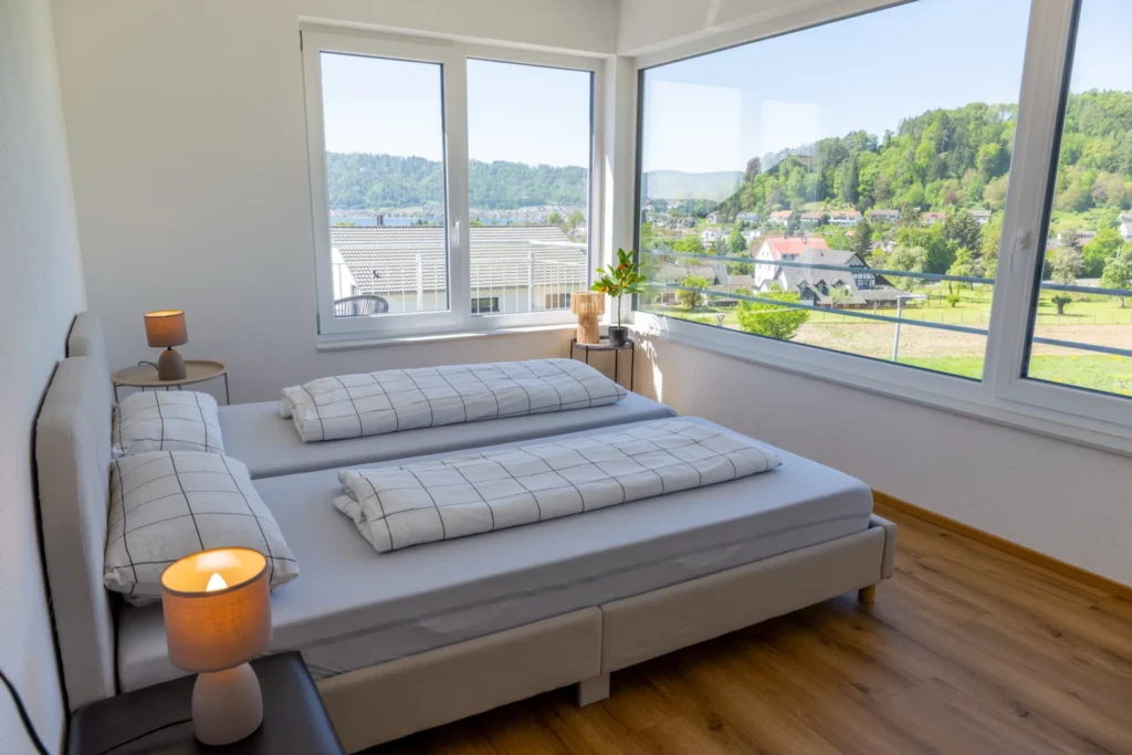 ferienhaus-aura-schlafzimmer-einzelbetten-panorama Schlafzimmer im Ferienhaus Aura mit zwei Einzelbetten, karierter Bettwaesche und Eckfenstern mit See- und Bergpanorama