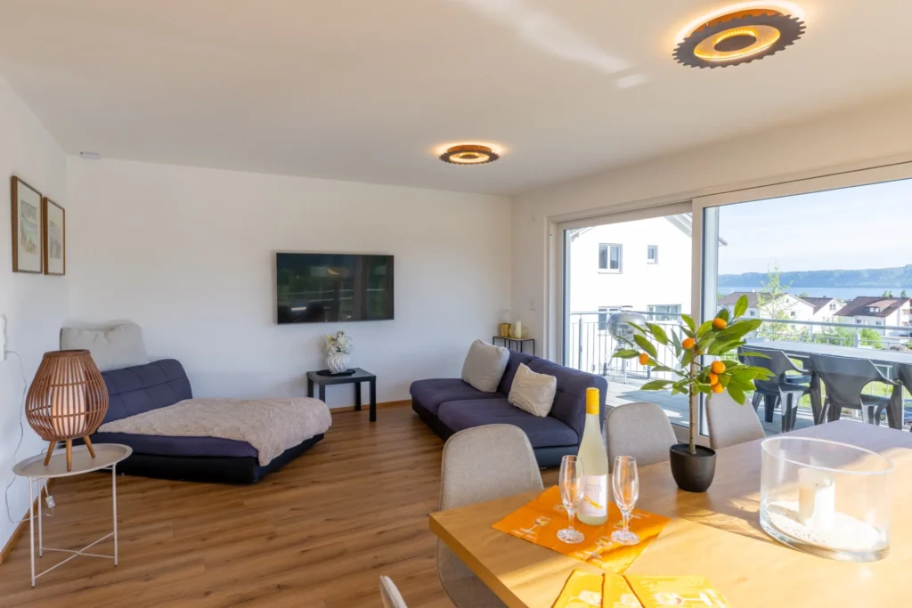 ferienhaus-aura-wohn-essbereich-seeblick-balkon Offener Wohn-Essbereich im Ferienhaus Aura mit Sofa, Esstisch, Weinglaesern und Balkon mit Seeblick