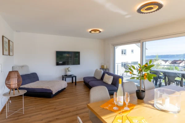 Offener Wohn-Essbereich im Ferienhaus Aura mit Sofa, Esstisch, Weinglaesern und Balkon mit Seeblick