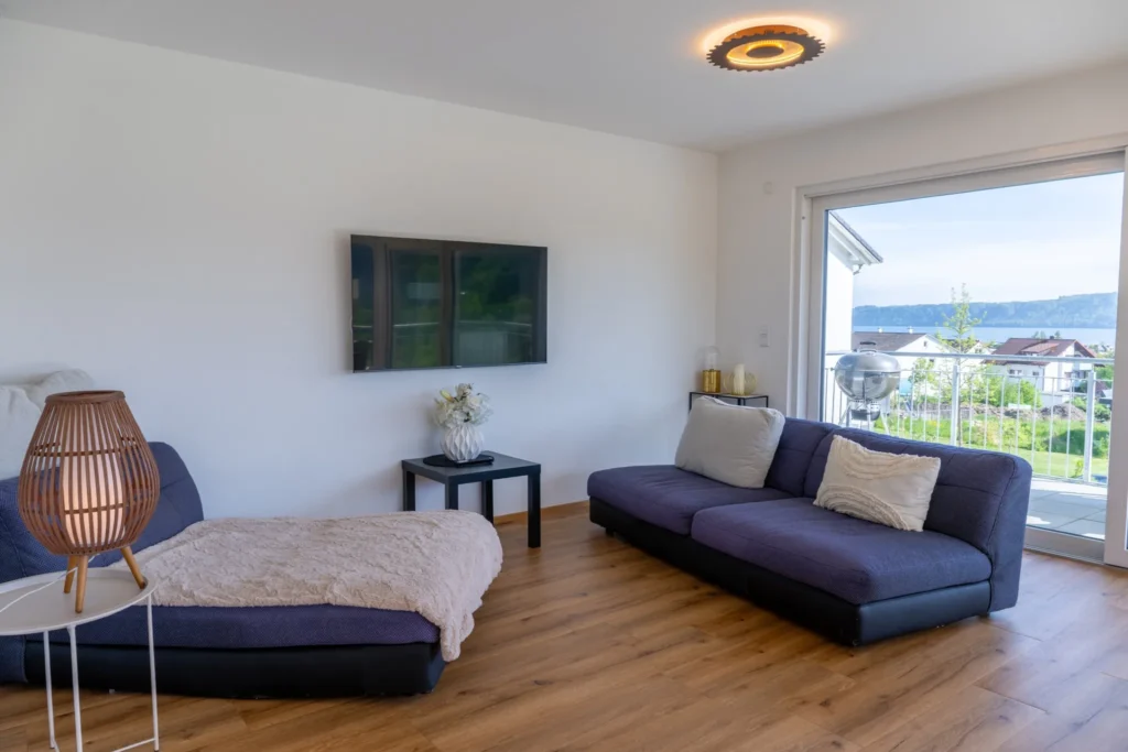 ferienhaus-aura-wohnzimmer-sofa-tv-seeblick Helles Wohnzimmer im Ferienhaus Aura mit blauem Sofa, Flachbild-TV und Balkontuer mit Bodensee-Blick
